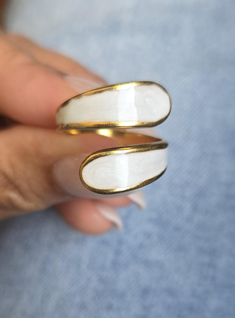 Puede incluir: Un anillo dorado con un dise&ntilde;o de esmalte blanco. El anillo se envuelve alrededor del dedo con dos secciones ovaladas de esmalte blanco, cada una delineada con oro. La pieza de joyer&iacute;a es un dise&ntilde;o moderno y minimalista.