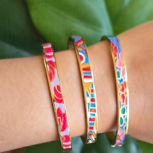 May include: Three gold-toned bangles with colorful enamel designs. The first bangle features red and purple floral patterns, the second has a geometric design with blue, orange, and red, and the third displays a floral pattern with pink and blue.
