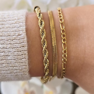 May include: Three gold bracelets of different designs are displayed on a wrist. The bracelets include a rope chain, a flat herringbone chain, and a curb chain. The background is blurred with white flowers.