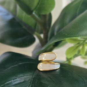 Puede incluir: Un anillo dorado con un dise&ntilde;o de esmalte blanco. El anillo tiene un dise&ntilde;o abierto &uacute;nico con dos secciones superpuestas. El anillo est&aacute; colocado sobre un fondo de hojas verdes, creando un contraste de colores y texturas.
