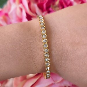 May include: Gold-toned bracelet with a series of clear, round gemstones set in a hexagonal pattern. The bracelet is worn on a wrist, with pink flowers in the background. The bracelet is a delicate piece of jewelry.