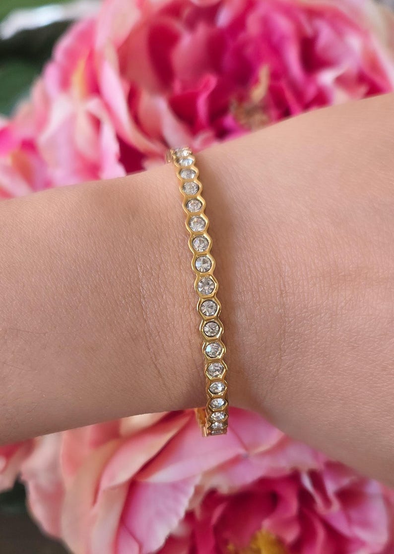 May include: Gold-toned bracelet with a series of clear, round gemstones set in a hexagonal pattern. The bracelet is worn on a wrist, with pink flowers in the background. The bracelet is a delicate piece of jewelry.