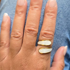 Puede incluir: Un anillo dorado con una incrustaci&oacute;n de esmalte blanco se lleva en un dedo. El anillo tiene un dise&ntilde;o envolvente. El fondo es una tela vaquera.