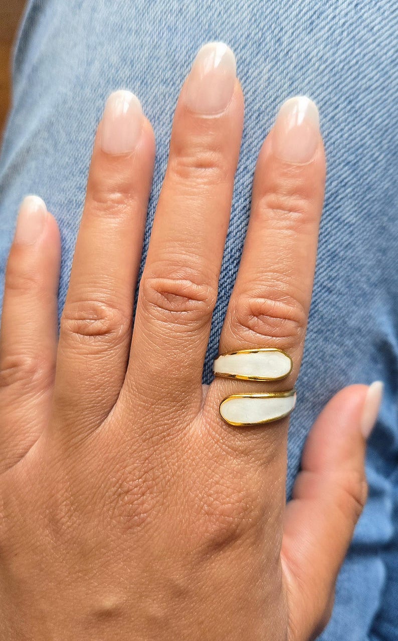 Puede incluir: Un anillo dorado con una incrustaci&oacute;n de esmalte blanco se lleva en un dedo. El anillo tiene un dise&ntilde;o envolvente. El fondo es una tela vaquera.
