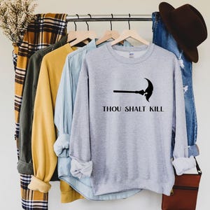 May include: A light gray sweatshirt with the text "THOU SHALT KILL" and a black scythe graphic. The sweatshirt is hanging on a clothing rack with other shirts and a pair of jeans.