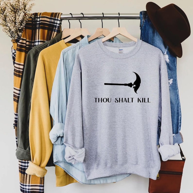 May include: A light gray sweatshirt with the text "THOU SHALT KILL" and a black scythe graphic. The sweatshirt is hanging on a clothing rack with other shirts and a pair of jeans.