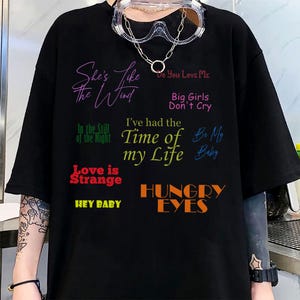 May include: Black t-shirt with multiple song titles in various colors. The phrases include "She's Like the Wind," "Do You Love Me," "Big Girls Don't Cry," "I've had the Time of my Life," and "Hungry Eyes."