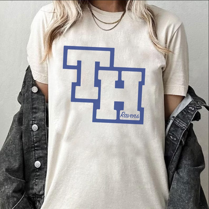 Peut inclure: T-shirt cr&egrave;me avec un motif bleu des lettres "TH" et le mot "Ravens" en dessous. Le t-shirt est port&eacute; avec une veste en jean et un collier dor&eacute;.