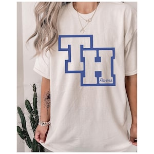 Peut inclure: T-shirt de couleur cr&egrave;me avec un motif bleu des lettres "TH" avec un contour bleu. Le mot "Ravens" est imprim&eacute; en bleu sous le "H". Le t-shirt est de style d&eacute;contract&eacute;.