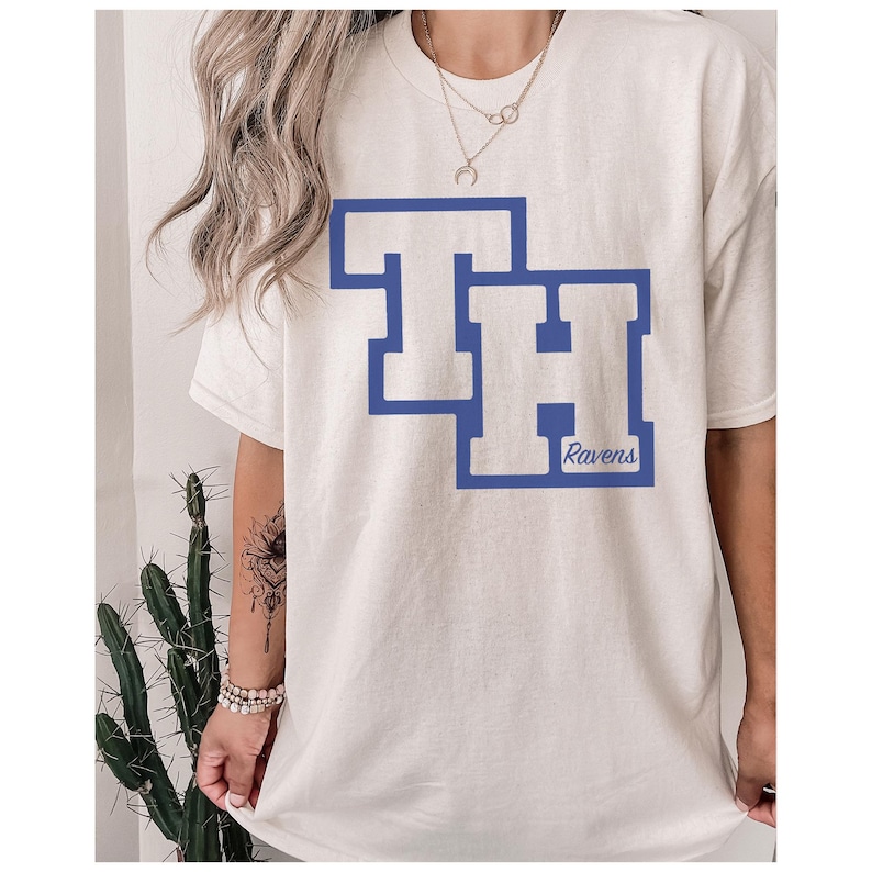 Peut inclure: T-shirt de couleur cr&egrave;me avec un motif bleu des lettres "TH" avec un contour bleu. Le mot "Ravens" est imprim&eacute; en bleu sous le "H". Le t-shirt est de style d&eacute;contract&eacute;.