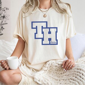 Peut inclure: T-shirt cr&egrave;me avec un motif bleu et blanc. Le motif pr&eacute;sente les lettres "TH" dans un style de lettres en bloc, avec le mot "Ravens" en dessous. Le t-shirt est de style d&eacute;contract&eacute;.