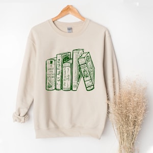 May include: Beige sweatshirt with a green graphic of stacked books. The book titles include "A Guest for Master Spider", "Ex Altiora", and "The Beginner's Tale". The sweatshirt is hanging on a wooden hanger.