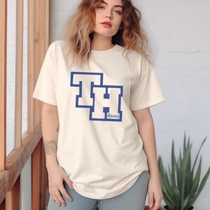 Peut inclure: T-shirt cr&egrave;me avec un motif bleu et blanc. Le motif pr&eacute;sente les lettres "TH" superpos&eacute;es, avec "Ravens" en dessous. Le t-shirt est de style d&eacute;contract&eacute;, &agrave; manches courtes.