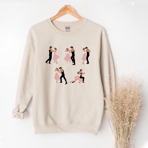 May include: Beige sweatshirt featuring illustrations of couples dancing in various poses. The dancers are depicted in black and pink, with the sweatshirt hanging on a wooden hanger. The design is repeated across the front of the garment.