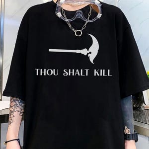 May include: Black t-shirt with a white graphic of a scythe and the text "THOU SHALT KILL". The shirt has short sleeves and a relaxed fit. A pair of clear safety glasses and a necklace are worn.