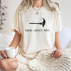 May include: Cream-colored t-shirt featuring a black graphic of a polearm and the text "THOU SHALT KILL" in a bold, sans-serif font. The shirt is made of a soft material and has short sleeves.