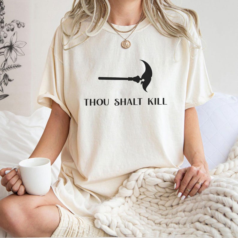 May include: Cream-colored t-shirt featuring a black graphic of a polearm and the text "THOU SHALT KILL" in a bold, sans-serif font. The shirt is made of a soft material and has short sleeves.