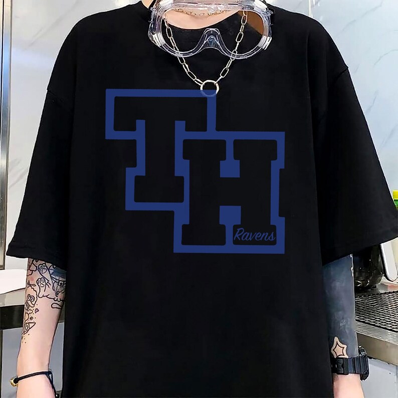 Peut inclure: T-shirt noir &agrave; manches courtes avec un motif graphique bleu et blanc. Le motif pr&eacute;sente les lettres "TH" dans un style universitaire, avec le mot "Ravens" en dessous. Le t-shirt est en mati&egrave;re souple.