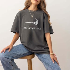May include: Dark grey t-shirt with the text "THOU SHALT KILL" and a graphic of a scythe. The shirt is a relaxed fit and is paired with light blue jeans. The model is sitting on a wooden stool.