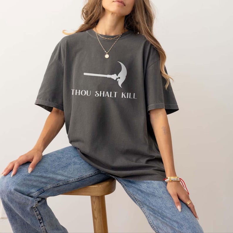 May include: Dark grey t-shirt with the text "THOU SHALT KILL" and a graphic of a scythe. The shirt is a relaxed fit and is paired with light blue jeans. The model is sitting on a wooden stool.
