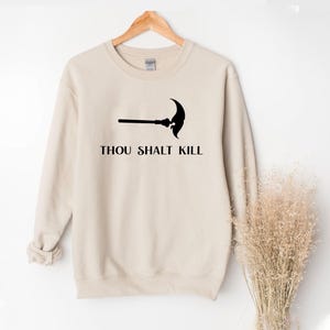 May include: A beige sweatshirt with a black graphic of a scythe and the text "THOU SHALT KILL" in a bold font. The sweatshirt is displayed on a wooden hanger.