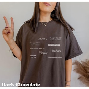 Może przedstawiać: Ciemnobrązowy t-shirt z białymi napisami. Frazy to m.in. "Mr. Spider doesn't like it", "Statement Begins" i "take her not me". Koszulka ma luźny krój.