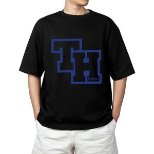 Peut inclure: T-shirt noir avec un motif bleu et noir des lettres "TH" et le mot "Ravens". Le t-shirt est &agrave; manches courtes et &agrave; col rond.