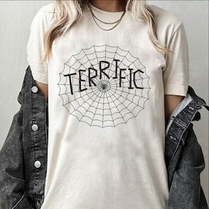 May include: Cream-colored t-shirt featuring a black spiderweb design with the word "TERRIFIC" in the center. The shirt is made of a soft material and has short sleeves. A dark denim jacket is worn over the shirt.