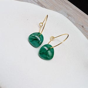 Peut inclure: Une paire de boucles d'oreilles créoles dorées avec des pendentifs verts marbrés en forme de goutte. Les boucles d'oreilles sont présentées sur une surface blanche, avec un fond en bois clair.