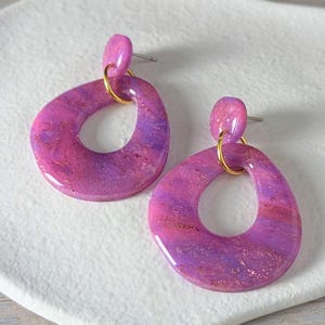 Könnte beinhalten: Ein Paar abstrakte Ohrringe in Rosa- und Lilatönen. Jeder Ohrring hat eine offene Tropfenform mit Goldglitzer. Die Ohrringe haben einen Stecker und einen kleinen goldfarbenen Ring, der den Tropfen mit dem Stecker verbindet.
