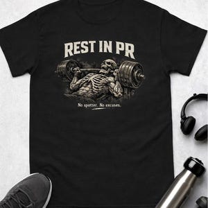 Puede incluir: Camiseta negra con un gráfico detallado de un esqueleto levantando una barra, con el texto "REST IN PR" y "No spotter. No excuses." El diseño tiene una estética vintage. La camiseta se muestra sobre una superficie clara.