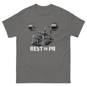 Puede incluir: Camiseta gris oscuro con un diseño de un esqueleto levantando pesas en un banco. El esqueleto está debajo de una barra con pesas. El texto "REST IN PR" está impreso en blanco debajo de la ilustración.