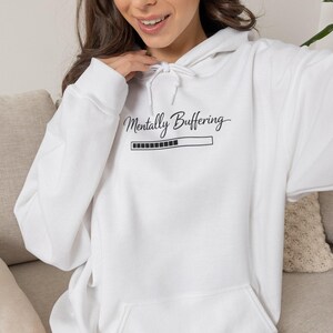 May include: A white hooded sweatshirt featuring a black graphic print on the chest that reads Mentally Buffering above a loading bar icon. The garment includes a drawstring hood, long sleeves, and a large front kangaroo pocket for storage.