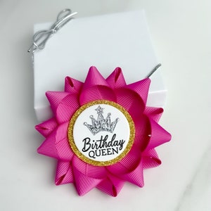 May include: A pink and gold birthday queen ribbon with a silver crown and glitter. The ribbon is tied into a bow and has a white circle with the text "Birthday Queen" in black.