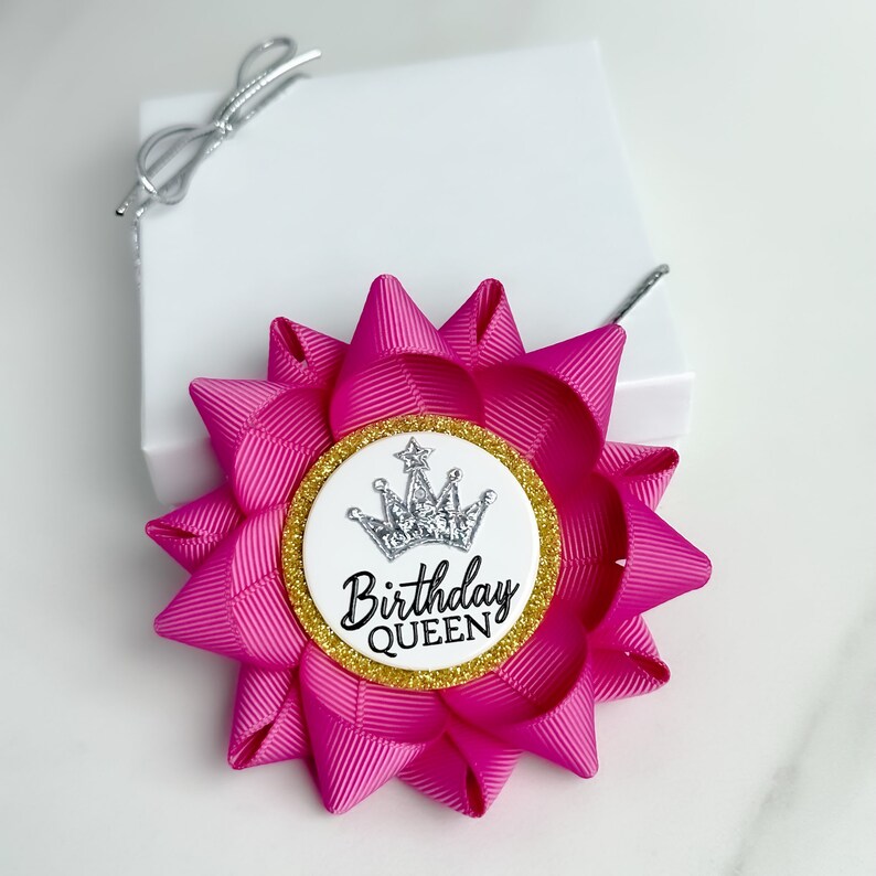 May include: A pink and gold birthday queen ribbon with a silver crown and glitter. The ribbon is tied into a bow and has a white circle with the text "Birthday Queen" in black.