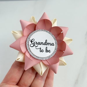 May include: A pink and cream bow with a white button that says "Grandma to be" in black script. The button has a silver glitter border.