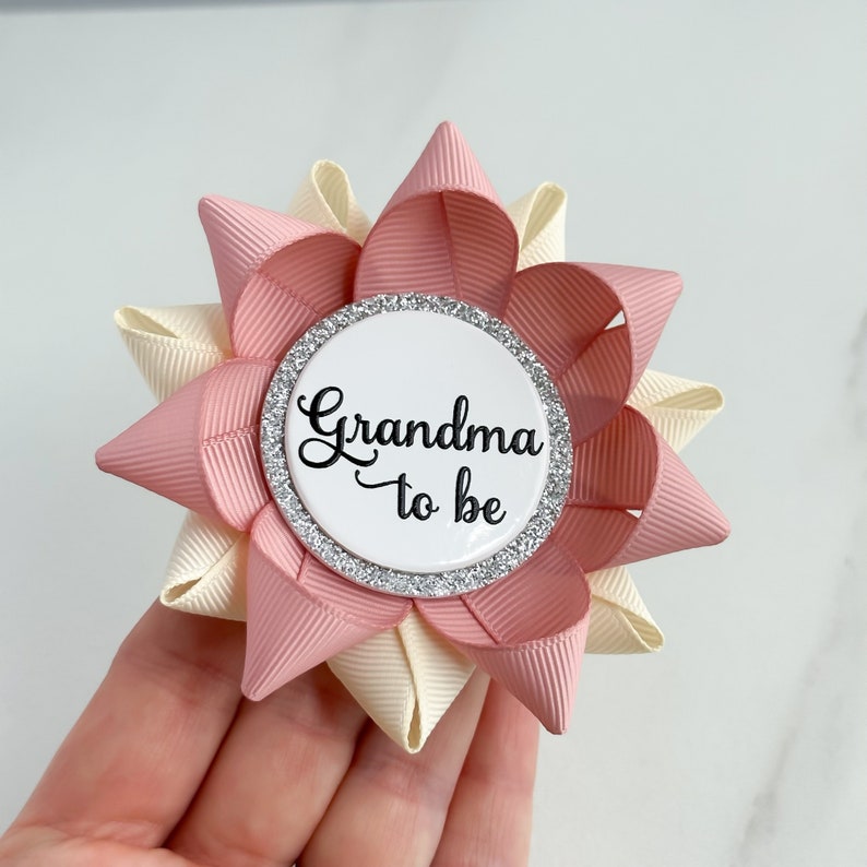 May include: A pink and cream bow with a white button that says "Grandma to be" in black script. The button has a silver glitter border.