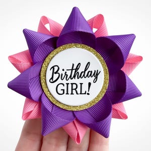May include: A decorative hair bow featuring a layered design with purple and pink ribbons. The center circle has gold glitter trim and the words "Birthday GIRL!" in black script. A festive accessory.