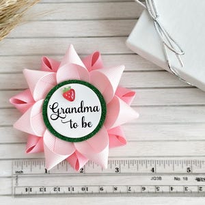 May include: Pink and green 'Grandma to be' button with a strawberry graphic. The button is attached to a pink bow.