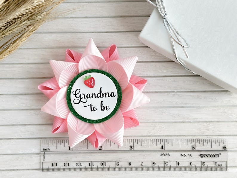 May include: Pink and green 'Grandma to be' button with a strawberry graphic. The button is attached to a pink bow.