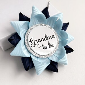 May include: A blue and navy blue bow with a white button that says "Grandma to be" in black script. The button has a silver glitter border.