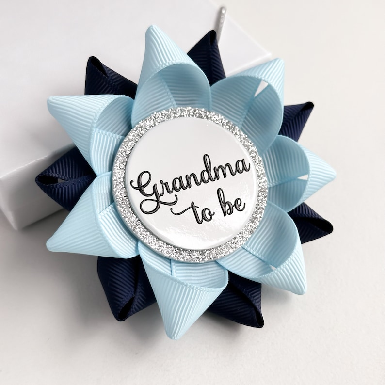May include: A blue and navy blue bow with a white button that says "Grandma to be" in black script. The button has a silver glitter border.
