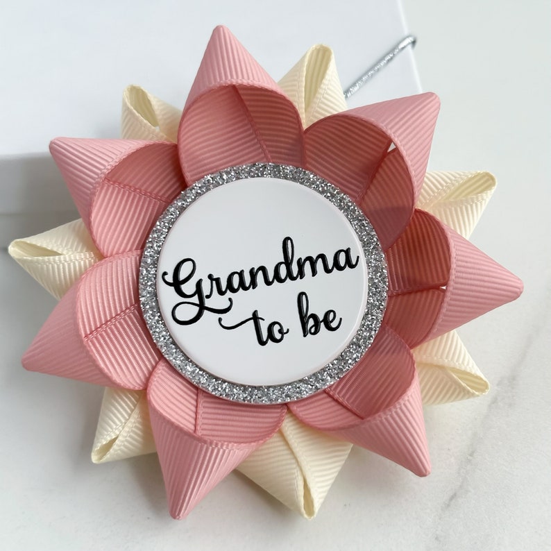 May include: A pink and cream flower-shaped pin with a white circle in the center that says "Grandma to be" in black script. The circle has a silver glitter border.