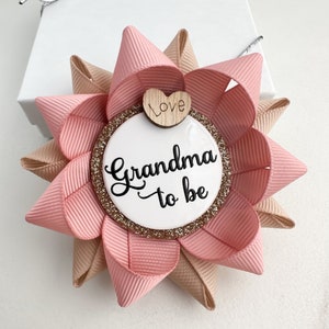 May include: A pink and beige bow with a white circle that says "Grandma to be" and a small wooden heart that says "Love".