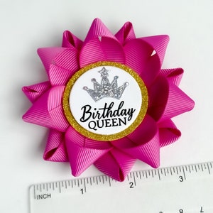 May include: Pink birthday bow with a white circle and gold glitter trim. The circle has a silver crown and the text "Birthday Queen".