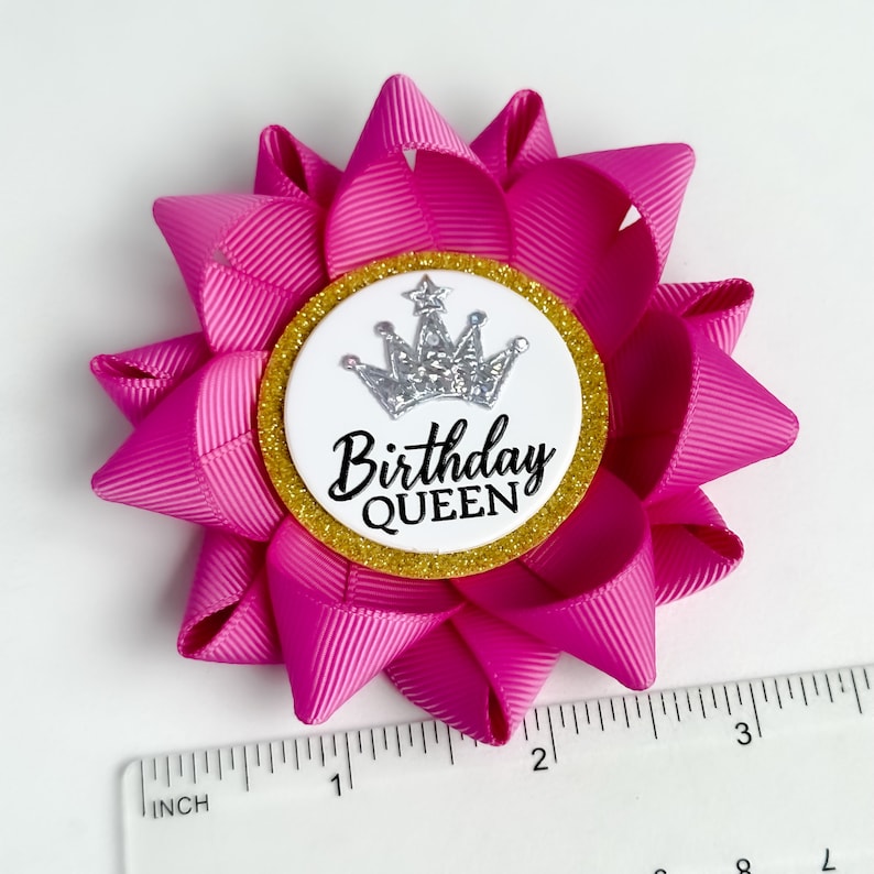 May include: Pink birthday bow with a white circle and gold glitter trim. The circle has a silver crown and the text "Birthday Queen".