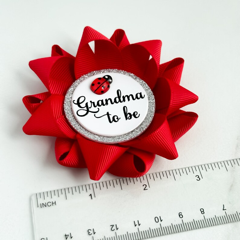 May include: Red and white grandma to be pin with a ladybug on a silver glitter background.