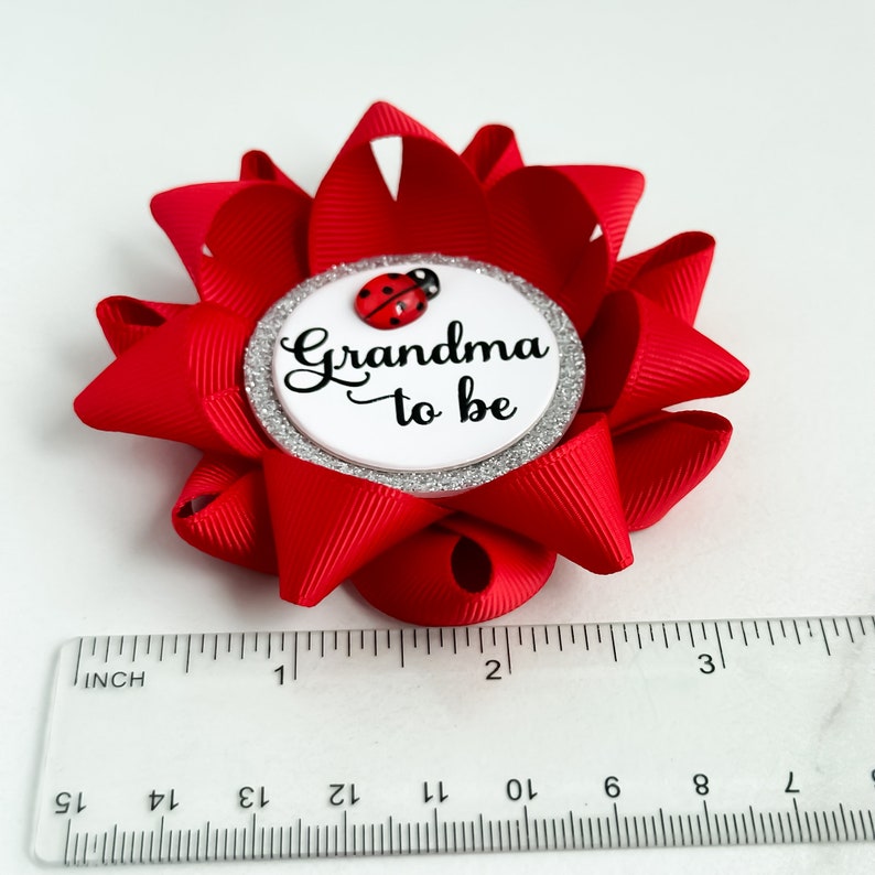 May include: Red and white 'Grandma to be' pin with a ladybug on the front. The pin is made of a red ribbon and has a white circle with silver glitter around the edge.