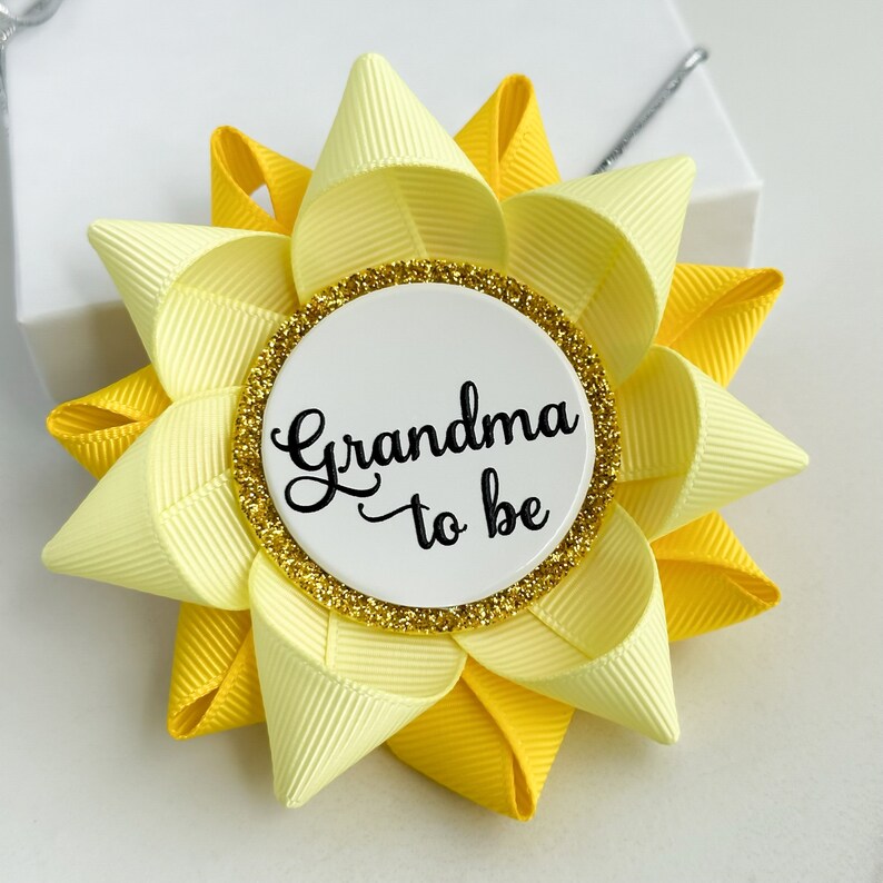 K&ouml;nnte beinhalten: Gelb und hellgelbe, blumenf&ouml;rmige Anstecknadel mit einem wei&szlig;en Kreis in der Mitte, auf dem in schwarzer Kursivschrift "Oma to be" steht. Der Kreis hat einen goldenen Glitzerrand.