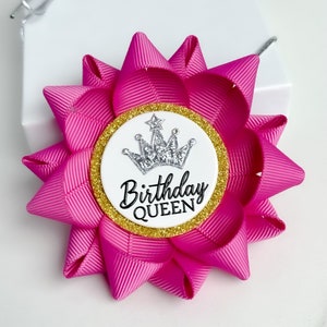 May include: A pink fabric bow with a white circle in the center. The circle has a silver crown with a star on top and the text "Birthday QUEEN" in black.